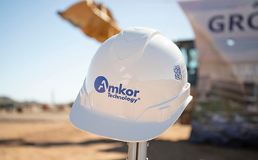 Close-up of a white Amkor Technology hard hat at the Peoria, Arizona groundbreaking site, with construction equipment and event signage blurred in the background.