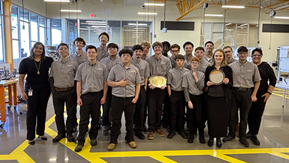 Group photo of Amkor team members and West-MEC students standing together in a modern manufacturing lab, with students in matching gray uniforms holding semiconductor wafers and awards in the center of the group.